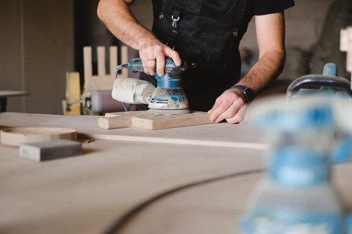 A person sanding wood with an electric sander in a workshop.