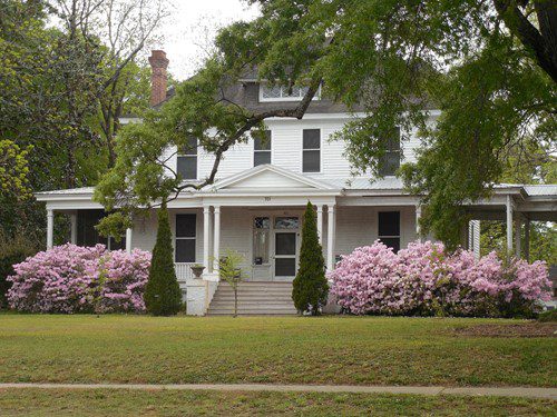 White two-story house with a porch and blooming pink azaleas.