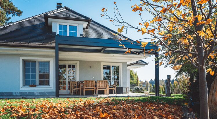 Modern house porch with wooden furniture and autumn leaves in the foreground.