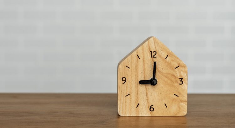 Wooden clock shaped like a house showing 3 o'clock on a wooden surface.