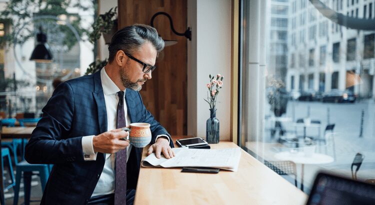 Man in suit reading newspaper and drinking coffee by window.