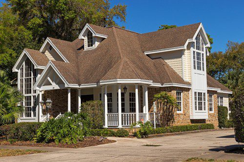 A charming suburban house with a wraparound porch and brown roof.
