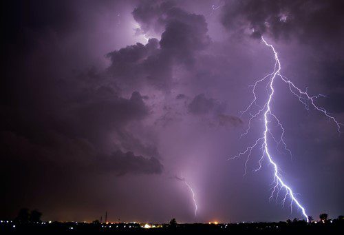 Striking lightning bolts illuminate a dark, stormy sky over a cityscape at night.