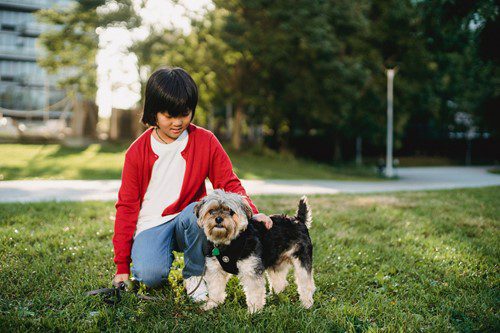 A woman in a red jacket kneeling next to her small dog outdoors.