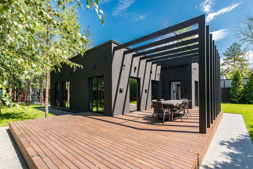 Modern black house with a spacious wooden deck and outdoor dining area under a pergola.