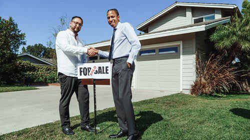 Two men shaking hands in front of a house with a for sale sign.