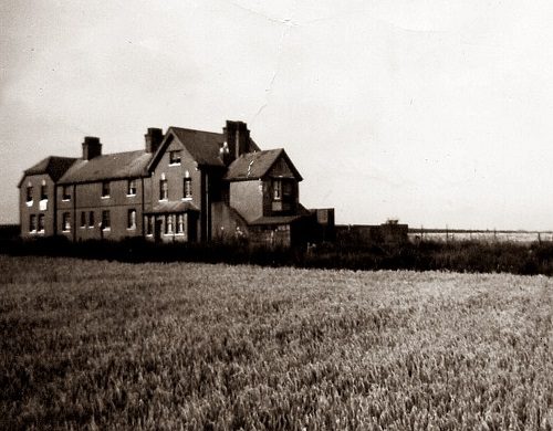 Old rural house beside an open field under a cloudy sky.
