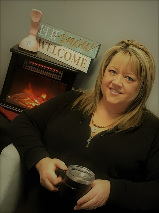 profile-21ad01e3-a38c-4cae-be76-0e8be17894b7 Smiling woman holding a drink, sitting near a cozy fireplace with a welcome sign.